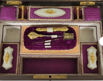 May include: An antique sewing kit with a dark wooden exterior and a plush purple interior. The kit contains gold-toned sewing tools, including a thimble, needle holder, and other ornate accessories. Decorative white and gold accents are also present.