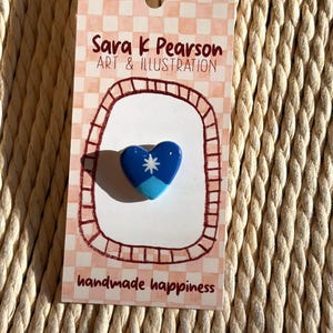 May include: A blue heart-shaped pin with a white star and light blue accents, displayed on a pink and white patterned card. The card reads "Sara k Pearson ART & ILLUSTRATION" and "handmade happiness."
