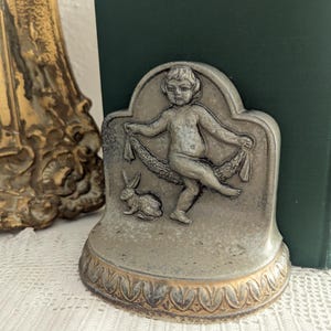 May include: A pair of decorative bookends made of metal with a gray and gold finish. The bookends feature a sculpted cherub holding a garland and a rabbit. The base of the bookends has a decorative leaf design.