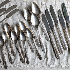 May include: An assortment of vintage silverware, including forks, spoons, and knives, arranged on a cream-colored tablecloth. The silverware has a shiny, silver finish and decorative detailing on the handles. A complete set for a formal dining experience.