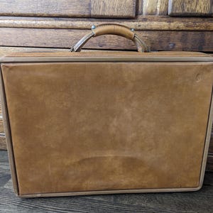 May include: A rectangular tan leather briefcase featuring a smooth front panel, a reinforced top handle with metallic hardware, and a structured frame. The briefcase has a minimalist design with clean edges and a subtle embossed logo on the right side.