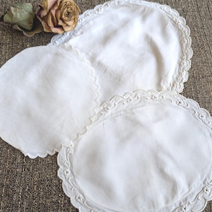May include: Three white, oval-shaped doilies with scalloped edges and delicate embroidered floral details. A dried rose with a brown and pink hue rests near the doilies. The doilies are arranged on a textured, brown surface.