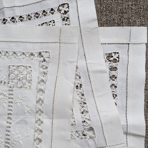 May include: Close-up of white linen textiles with intricate cutwork and embroidered floral and geometric designs. The textiles feature delicate openwork patterns and decorative stitching along the edges, showcasing a vintage aesthetic.