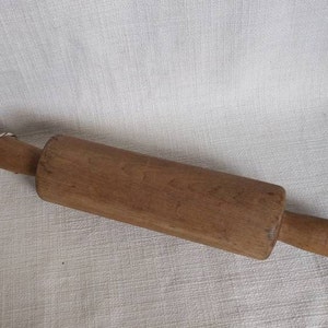 May include: A wooden rolling pin with a cylindrical body and tapered handles. The rolling pin is made of light brown wood and appears to be well-used. It is a kitchen utensil for rolling out dough.