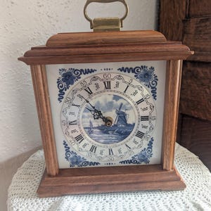 May include: A square wooden mantel clock with a brass handle on top. The clock face features a white background with blue floral corner patterns, black Roman numerals, and a central circular illustration of a windmill by a river.
