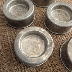 May include: A collection of five small, round, silver-colored metal containers. Each container has a shallow, circular interior and a slightly raised, rounded edge. The metal shows signs of wear, with some discoloration and scratches.