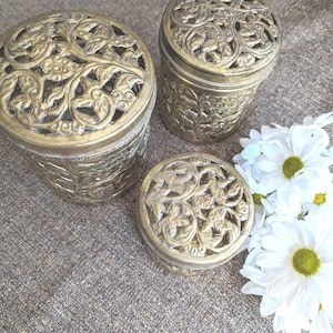 May include: Three antique brass containers with intricate floral cut-out designs. The containers are cylindrical and round, varying in size. White daisies with yellow centers are arranged to the right of the containers.