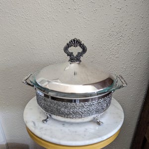 May include: Silver-plated chafing dish with a glass insert and ornate handle. The dish has a textured, patterned band around the base and sits on three decorative feet. The lid features a decorative handle. A classic serving piece.