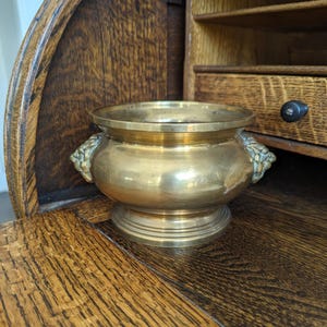 May include: A small, antique brass bowl with a wide rim and a rounded body. The bowl features two lion head handles and sits on a circular base. The bowl is set against a backdrop of a wooden roll-top desk.