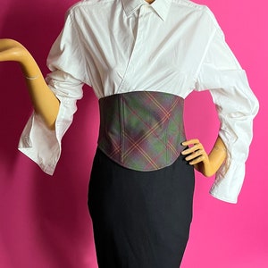 May include: A white button-down shirt with a plaid corset belt and a black pencil skirt. The mannequin is standing on a black octagonal base.