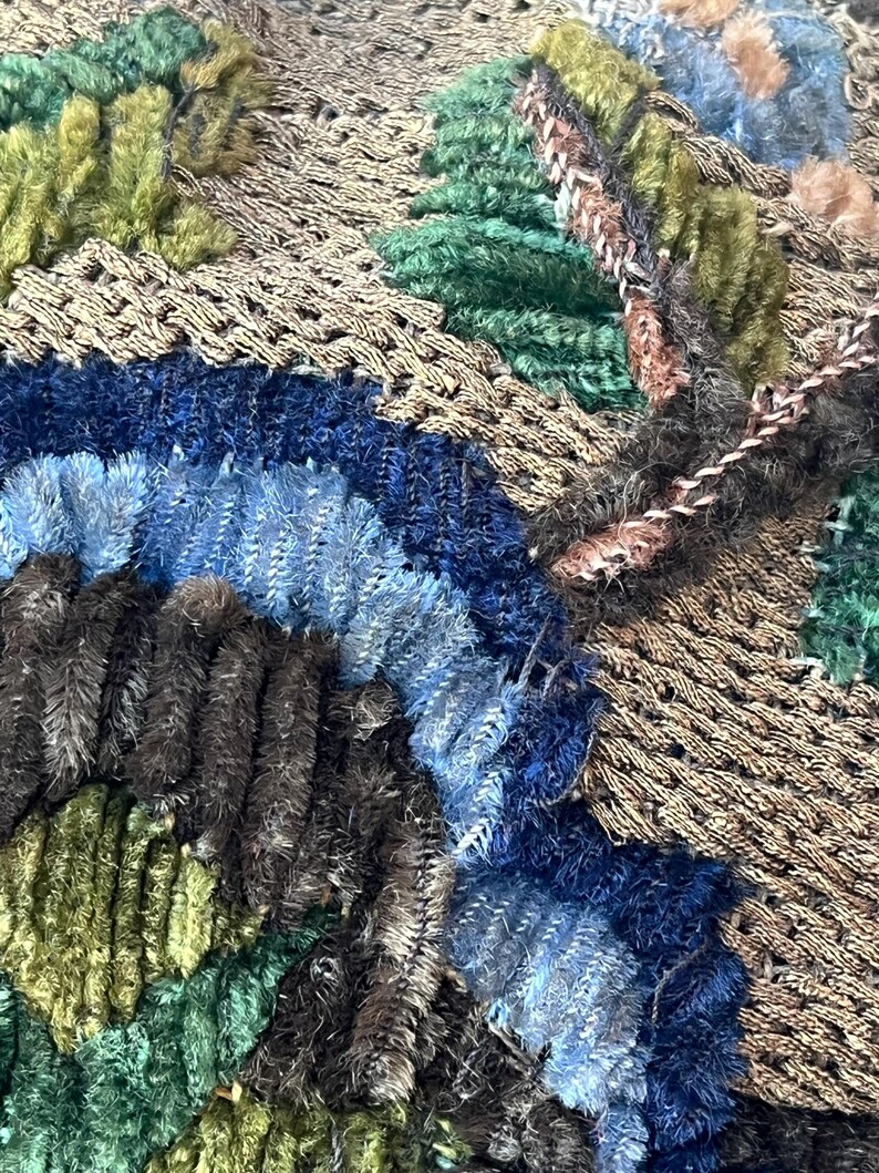 May include: Close-up of a brown fabric with blue, green, and brown embroidery. The fabric has a textured, woven appearance.