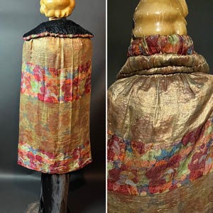May include: A golden, shimmering fabric garment with a black collar and floral accents. The garment has a cylindrical shape and is displayed on a dark wooden stand. The floral accents feature red, blue, and green hues.