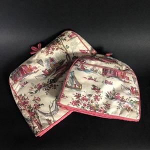 Könnte beinhalten: Zwei gesteppte Teewärmer mit einem cremefarbenen Toile-Muster mit rosa, roten und blauen Akzenten. Das Muster zeigt Szenen mit Menschen, Booten und Bäumen. Die Kanten sind mit rosa Paspeln und einer rosa Schleife versehen.