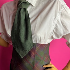 May include: A white button-down shirt with a dark green tie knotted at the collar. The shirt is worn over a plaid corset with a brown, green, and red pattern.