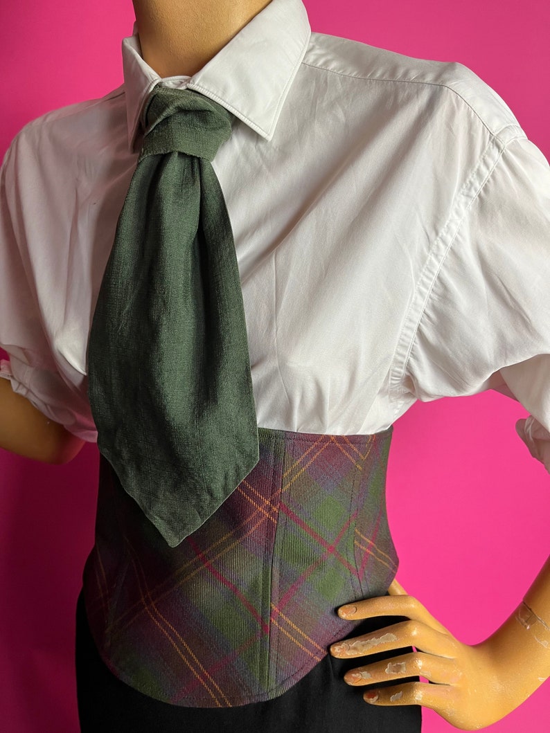 May include: A white button-down shirt with a dark green tie knotted at the collar. The shirt is worn over a plaid corset with a brown, green, and red pattern.