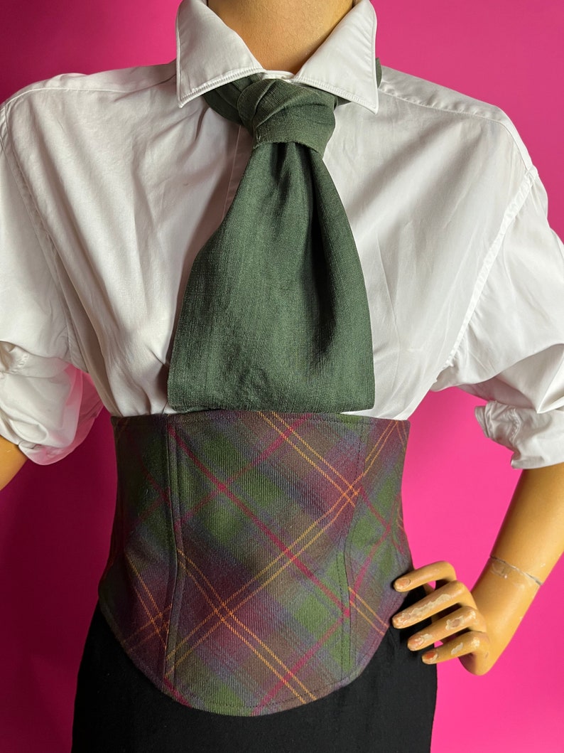 May include: A white button-down shirt with a green linen tie and a plaid corset. The corset has a green, red, and brown plaid pattern.