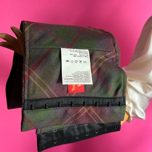 May include: A black and green plaid corset with black hook and eye closures. The corset has a red label with the word "Vivi" on it. The label also includes care instructions for washing and drying the garment.