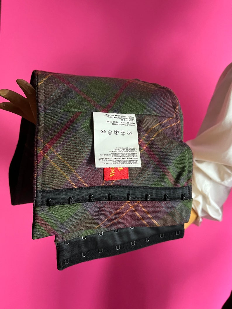 May include: A black and green plaid corset with black hook and eye closures. The corset has a red label with the word "Vivi" on it. The label also includes care instructions for washing and drying the garment.
