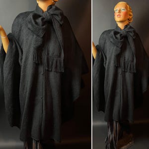 Op de afbeelding: Een donkergrijze cape met een grote strik bij de hals en een sjaal met franjes. De cape heeft een knoopsluiting aan de voorkant en wordt getoond op een mannequin. Het kledingstuk lijkt gemaakt van een zacht, getextureerd materiaal.