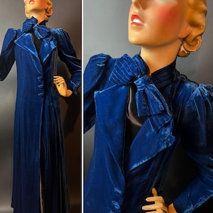 May include: A long, royal blue velvet coat with a bow detail at the neck and puffed sleeves. The coat has a button-down front and a tailored fit. The vintage coat is displayed on a mannequin.