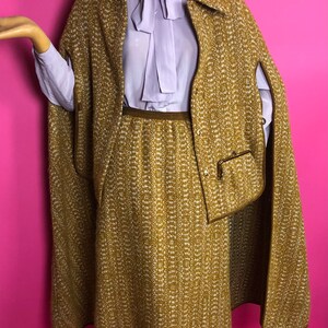 May include: A brown and white tweed skirt and matching cape with a brown trim. The skirt is knee-length and the cape has a collar and a pocket.