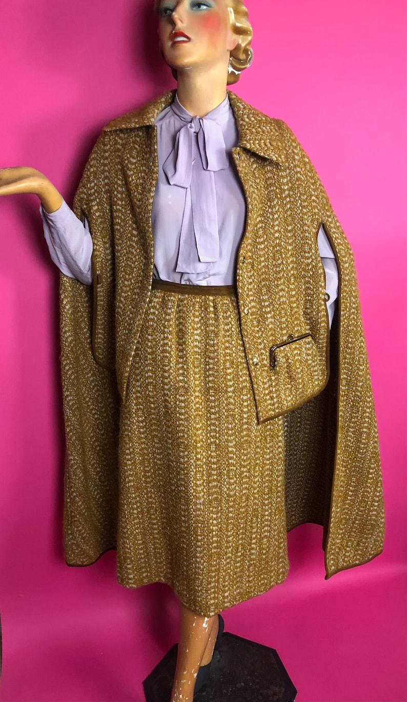 May include: A brown and white tweed skirt and matching cape with a brown trim. The skirt is knee-length and the cape has a collar and a pocket.