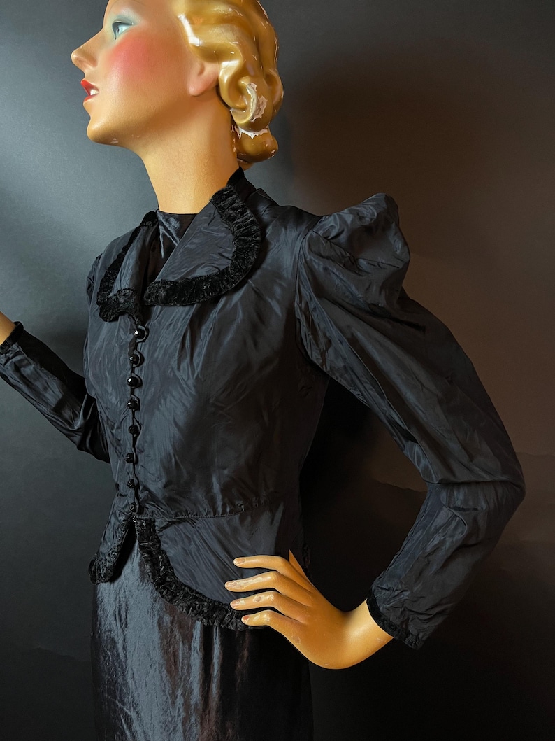 Op de afbeelding: Een zwarte vintage jurk met een getailleerd bovenstuk en een volle rok. De jurk heeft een hoge kraag met een fluwelen rand en een rij knopen aan de voorkant. De mouwen zijn lang en gepoft aan de schouders.