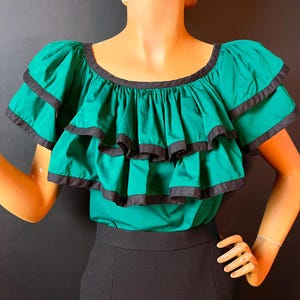 Könnte beinhalten: Ein grünes Oberteil mit Rüschen und schwarzem Besatz. Das Oberteil hat kurze Ärmel und einen runden Ausschnitt. Das Model trägt einen schwarzen Rock. Das Oberteil ist im Vintage-Stil.