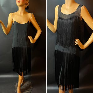 May include: A black vintage dress with a flapper style. The dress features a fitted bodice with small buttons and a fringed skirt. The dress is displayed on a mannequin.