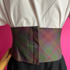 May include: A brown, green, and purple plaid corset belt with a brown leather-like finish. The belt has a hook and eye closure in the back.
