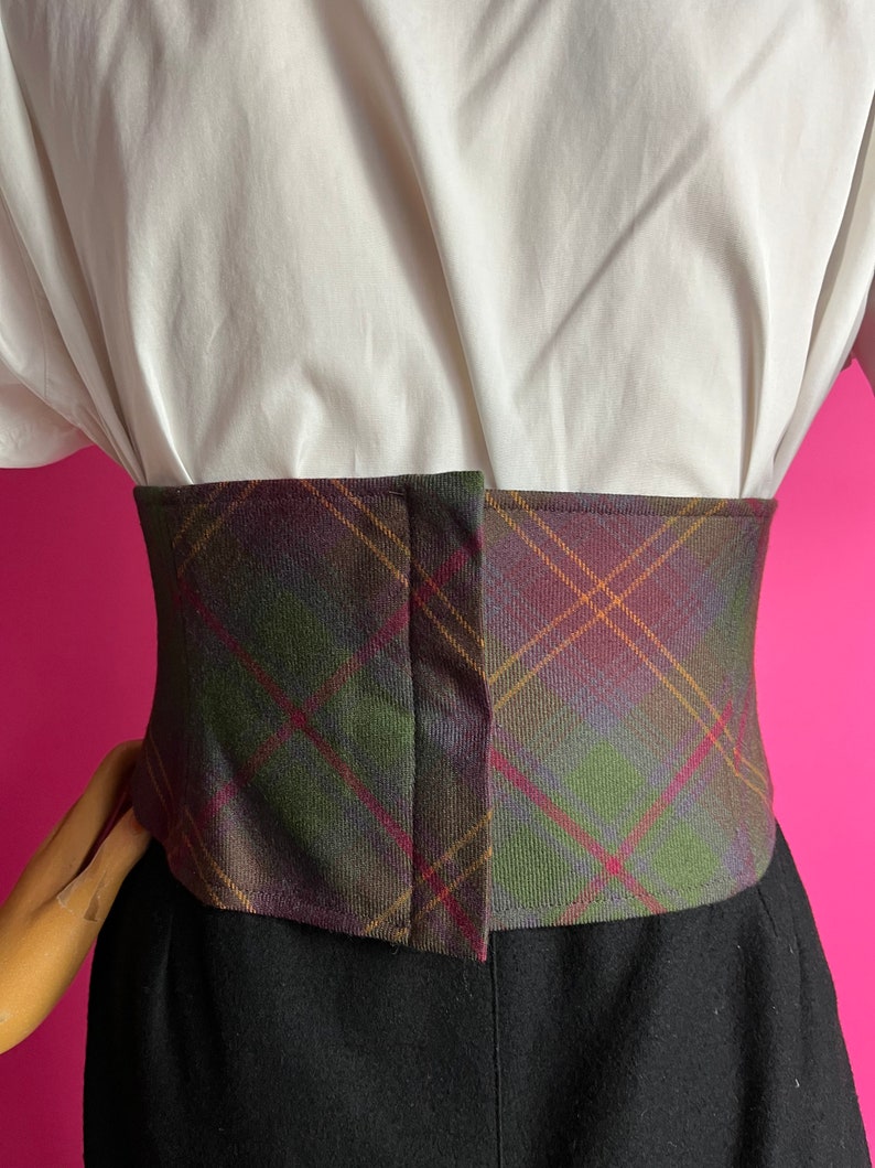 May include: A brown, green, and purple plaid corset belt with a brown leather-like finish. The belt has a hook and eye closure in the back.