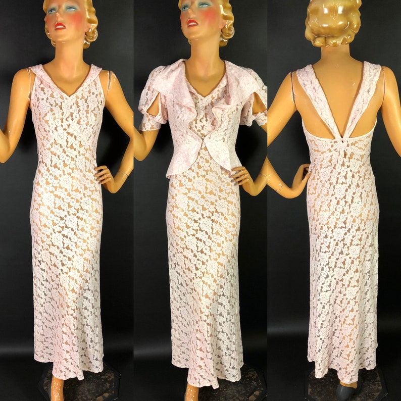 May include: A white lace dress with a V-neckline and a fitted bodice. The dress has a sheer overlay and a long, flowing skirt. The dress is shown on a mannequin.