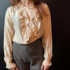 以下が含まれることがあります： 前面にフリルがあしらわれたクリーム色のブラウス。長袖トップスは、グレー、ブラウン、赤のチェック柄のツイードショーツと組み合わせています。モデルはラベンダー色のタイツを着用しています。