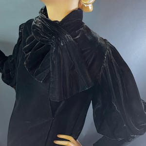 May include: A black velvet dress with a high ruffled collar and long, puffed sleeves. The dress is displayed on a mannequin, showcasing its vintage design and elegant silhouette. The fabric appears soft and luxurious.