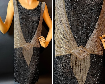 May include: A black beaded flapper dress with gold accents. The sleeveless dress features a scoop neckline and a geometric design with gold beading across the bodice and a beaded fringe detail.