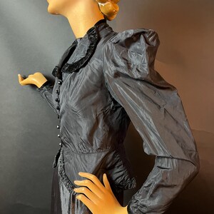 Op de afbeelding: Een zwarte zijden blouse met een fluwelen rand en pofmouwen. De blouse heeft een knoopsluiting aan de voorkant en een getailleerde pasvorm.