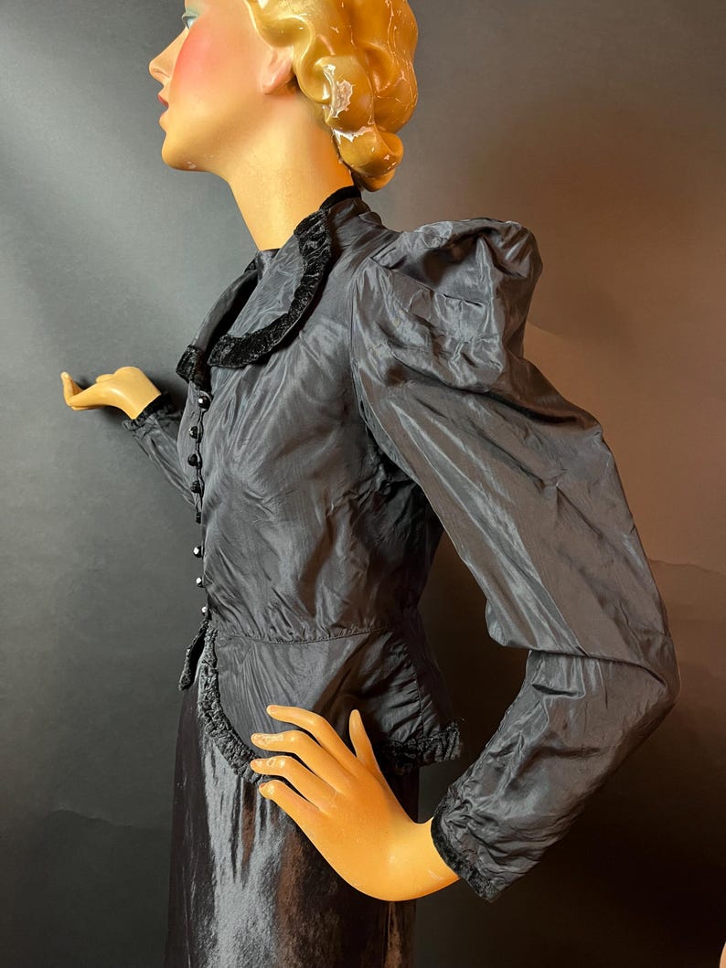 Op de afbeelding: Een zwarte zijden blouse met een fluwelen rand en pofmouwen. De blouse heeft een knoopsluiting aan de voorkant en een getailleerde pasvorm.