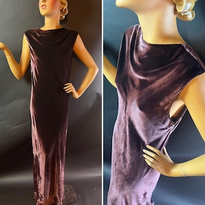 May include: A floor-length, sleeveless dress in a deep burgundy hue. The dress features a cowl neckline and a form-fitting silhouette, made of a soft, velvety fabric. The dress is displayed on a mannequin.