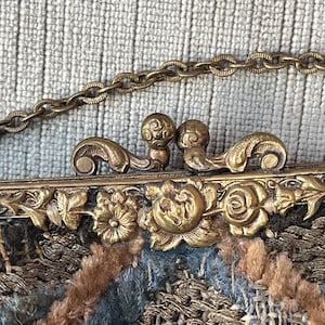 May include: A vintage embroidered purse with a gold metal clasp and chain. The purse is made of a variety of fabrics, including velvet, silk, and wool. The clasp is decorated with floral motifs.
