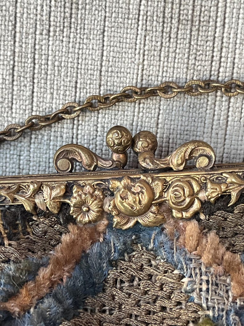 May include: A vintage embroidered purse with a gold metal clasp and chain. The purse is made of a variety of fabrics, including velvet, silk, and wool. The clasp is decorated with floral motifs.