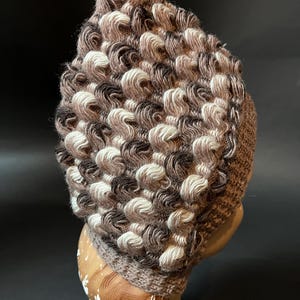May include: A hand-knitted beanie hat with a distinctive textured pattern. The hat combines brown, cream, and dark brown yarns, forming a visually appealing design. The hat is shown on a mannequin head.