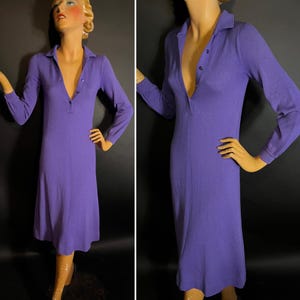 May include: A long-sleeved, knee-length purple dress with a collared neckline and a button-down placket. The dress is displayed on a mannequin against a black background. The dress has a fitted silhouette.
