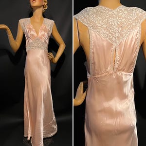 May include: A floor-length pale pink satin gown featuring a deep V-neckline and cap sleeves. The bodice is accented with intricate white floral lace panels at the shoulders and waist. The back displays a matching lace yoke and a vertical slit.