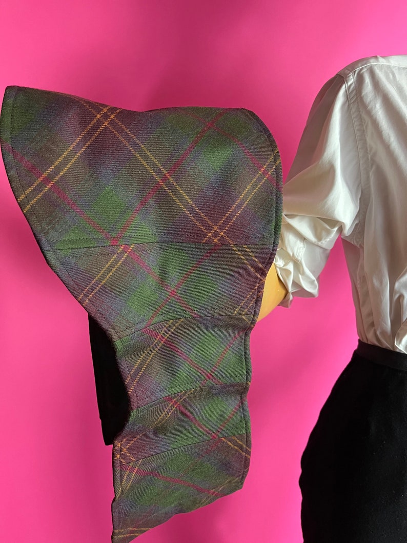 May include: A large, wide-brimmed hat made of a plaid fabric with green, purple, and brown tones. The hat is folded in half and has a curved shape.
