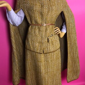 May include: A brown and white tweed cape and skirt set with a brown leather belt. The cape has a collar and a long, flowing design. The skirt is knee-length and has a straight silhouette.