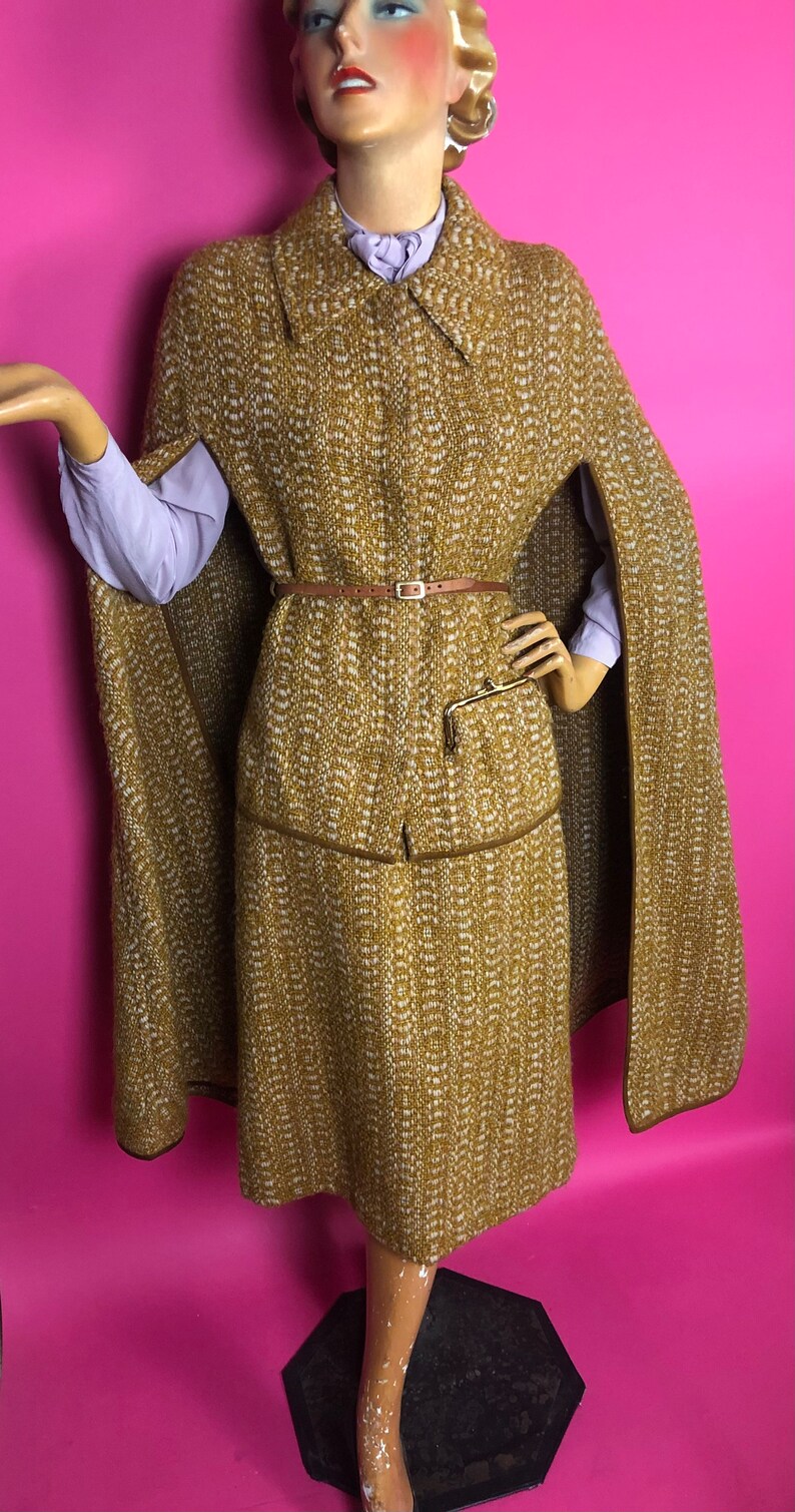 May include: A brown and white tweed cape and skirt set with a brown leather belt. The cape has a collar and a long, flowing design. The skirt is knee-length and has a straight silhouette.