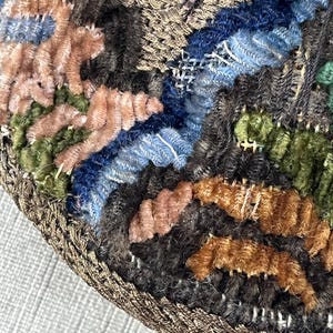 May include: Close-up of a circular, multi-colored, embroidered fabric with a brown, braided border. The fabric is made of different textures and colors, including pink, blue, green, and brown.