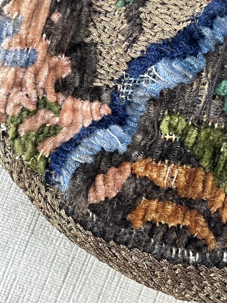 May include: Close-up of a circular, multi-colored, embroidered fabric with a brown, braided border. The fabric is made of different textures and colors, including pink, blue, green, and brown.