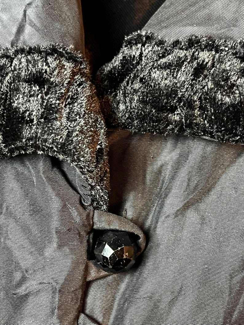 Op de afbeelding: Close-up van een zwarte fluwelen kraag met een zwarte knoop. De kraag is bevestigd aan een zwart kledingstuk.