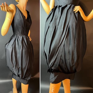 May include: A black sleeveless dress with a V-neck and a distinctive bubble skirt. The dress is made of a shiny material and has a fitted bodice. The skirt features vertical pleats, creating a voluminous shape. The dress is displayed on a mannequin.